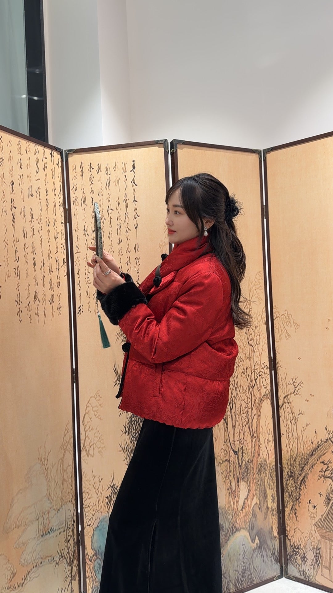 Woman in a red coat holding a pen in front of an ornate screen with artistic designs.