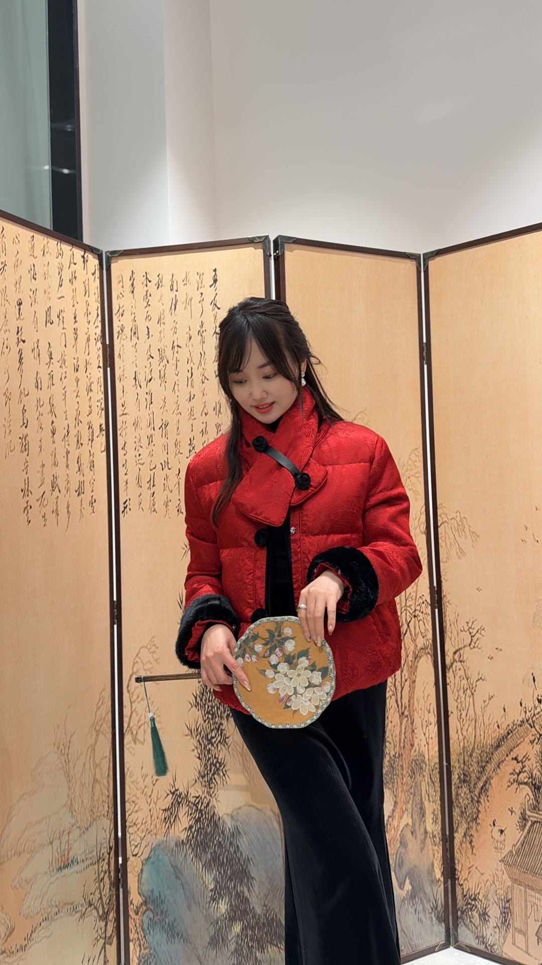 Woman in a red coat holding a round object in front of an ornate screen.